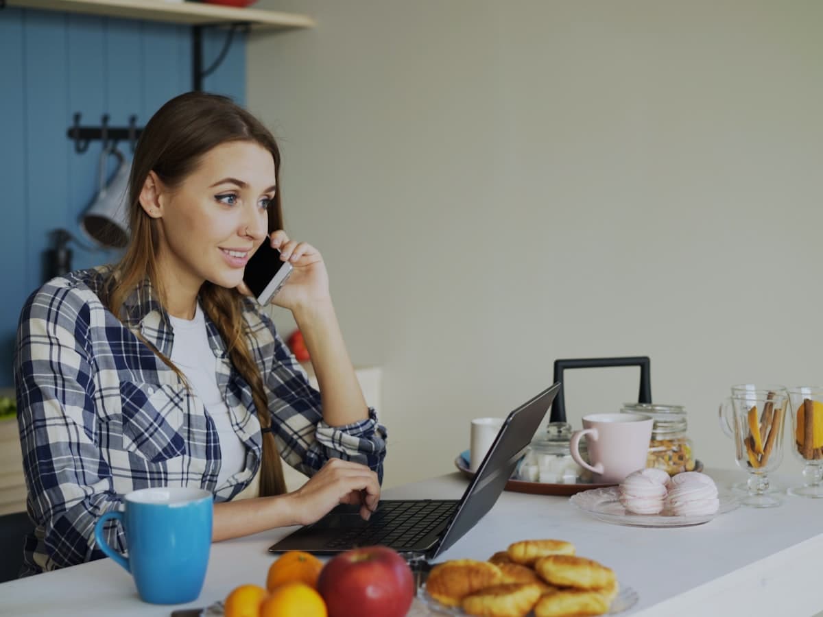 Someone at a kitchen table with a laptop and phone, sorting out paperwork